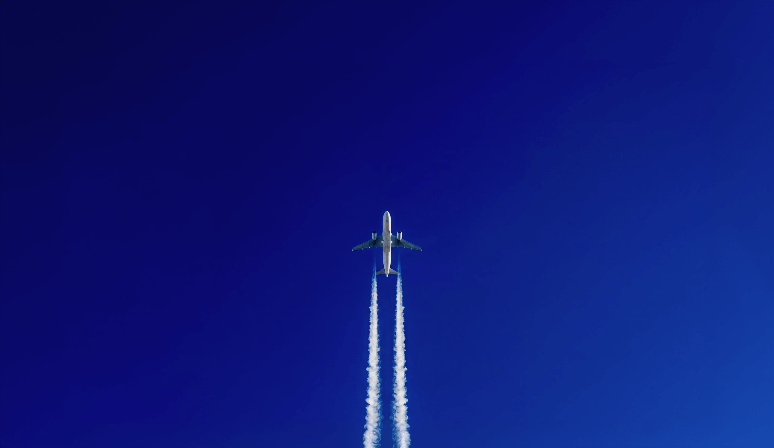 Airplane flying upward in clear blue sky leaving two vapor trails behind.