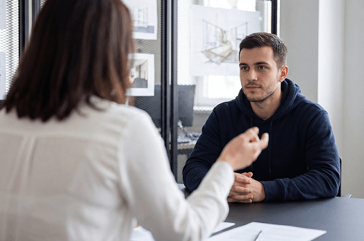 Man in navy hoodie attentively listens to a woman speaking across a table in an office.