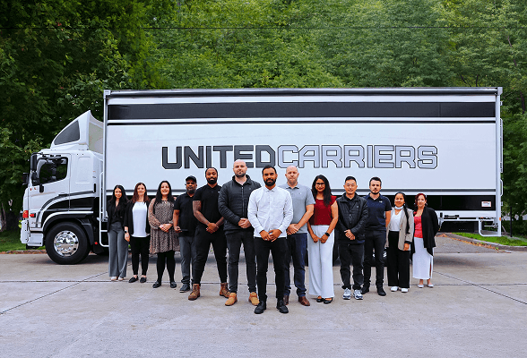 Group of twelve diverse people standing in front of a United Carriers truck outdoors.