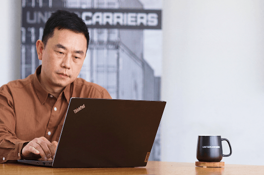 Man in brown shirt working on a ThinkPad laptop at a table with a black mug labeled United Carriers.